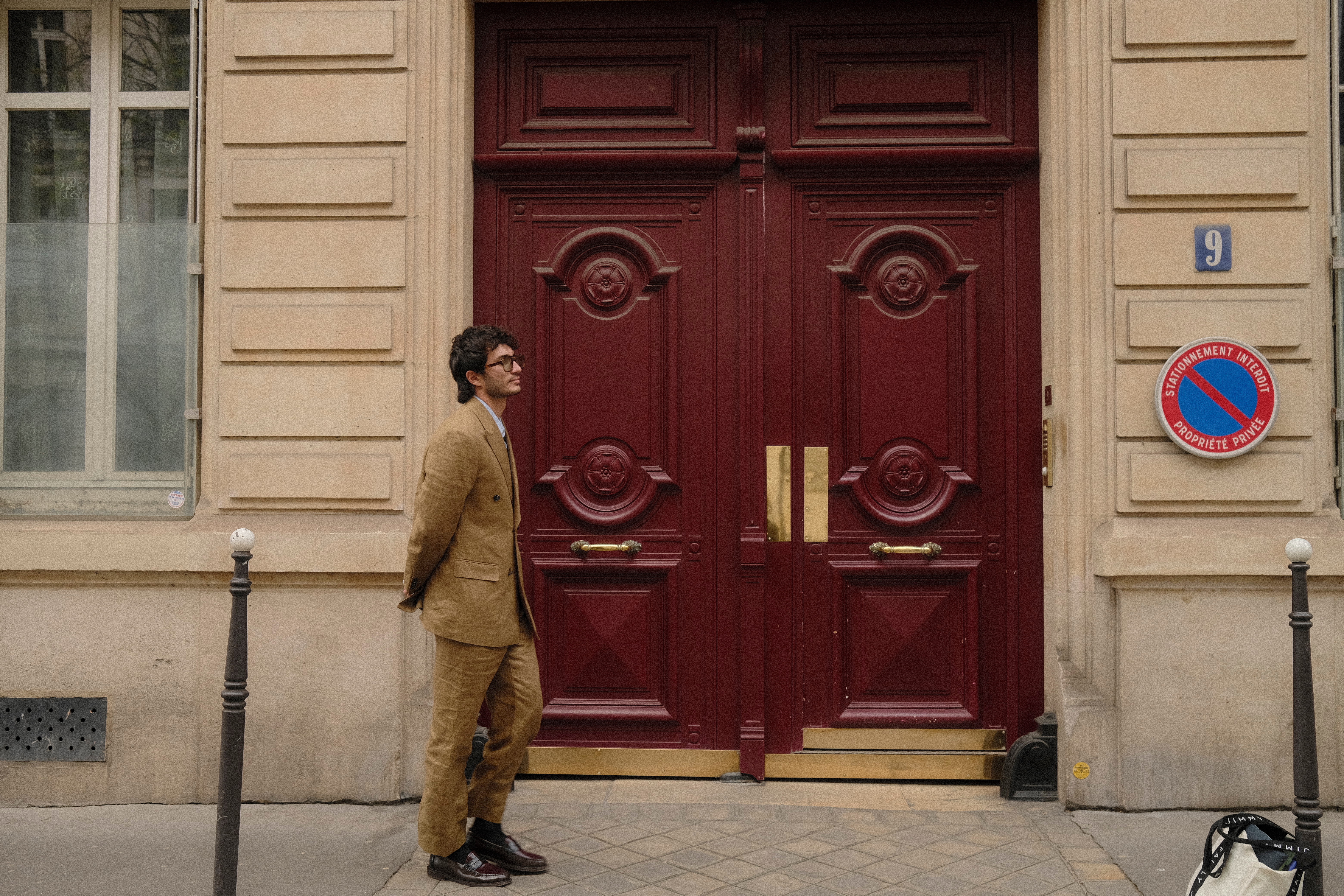 A man in a camel double-breasted Blitzer suit standing before deep red Parisian doors